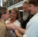 A military promotion or pinning ceremony is taking place, with a uniformed service member receiving insignia or rank pins on her collar. Two civilians — a man in a light blue shirt and a woman in white — are actively pinning the insignia, while a third person with a gray beard observes in the background. The setting appears to be an indoor military facility or hangar.