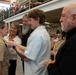 A military promotion or pinning ceremony is taking place in what appears to be a naval facility or hangar. A female service member in tan/khaki Navy uniform is having insignia pinned onto her collar by a civilian man in a light blue shirt, surrounded by family members. Uniformed naval personnel and spectators are visible in the background on bleachers.