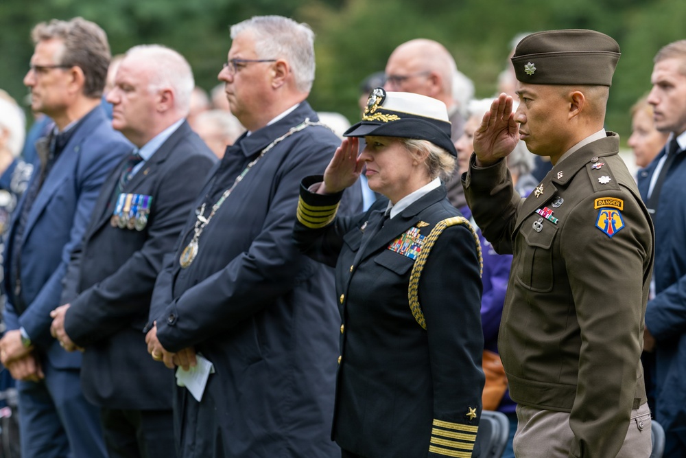 A formal military and civilian ceremony is taking place outdoors, likely a memorial or commemorative event. Two military officers in the foreground — one in U.S. Navy dress uniform and one in U.S. Army service uniform — are rendering a salute. Behind them stand civilians and other officials, some wearing medals and ceremonial chains of office.