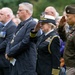 A formal military and civilian ceremony is taking place outdoors, likely a memorial or commemorative event. Two military officers in the foreground — one in U.S. Navy dress uniform and one in U.S. Army service uniform — are rendering a salute. Behind them stand civilians and other officials, some wearing medals and ceremonial chains of office.