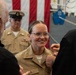 A U.S. Navy promotion or pinning ceremony is taking place, with a female sailor in khaki service uniform being pinned by two civilians, likely family members. An American flag is visible in the background along with a senior enlisted Navy officer in uniform observing the ceremony. The sailor is smiling broadly, indicating a celebratory milestone event.