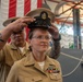 A U.S. Navy promotion or advancement ceremony is taking place, with one sailor placing a chief petty officer cover (hat) on another sailor's head. The recipient is a female sailor in khaki service uniform adorned with multiple ribbons, warfare devices, and chief petty officer collar insignia. A large American flag serves as the backdrop in what appears to be a hangar bay or similar military facility.