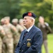 An elderly veteran stands at attention in a military cemetery, wearing a dark navy blazer with regimental insignia and a dark beret with a red hackle and gold badge. White Commonwealth War Graves Commission-style headstones are visible in the foreground and background. Soldiers in camouflage uniforms are visible out of focus in the background, suggesting a formal commemorative ceremony.
