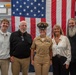 A group of five people pose for a formal photograph in front of a large American flag inside what appears to be a naval facility or museum. The central figure is a woman in U.S. Navy dress khaki uniform with a chief petty officer cover and multiple ribbons/medals, surrounded by four civilians. The setting and attire suggest a military promotion ceremony or official recognition event.