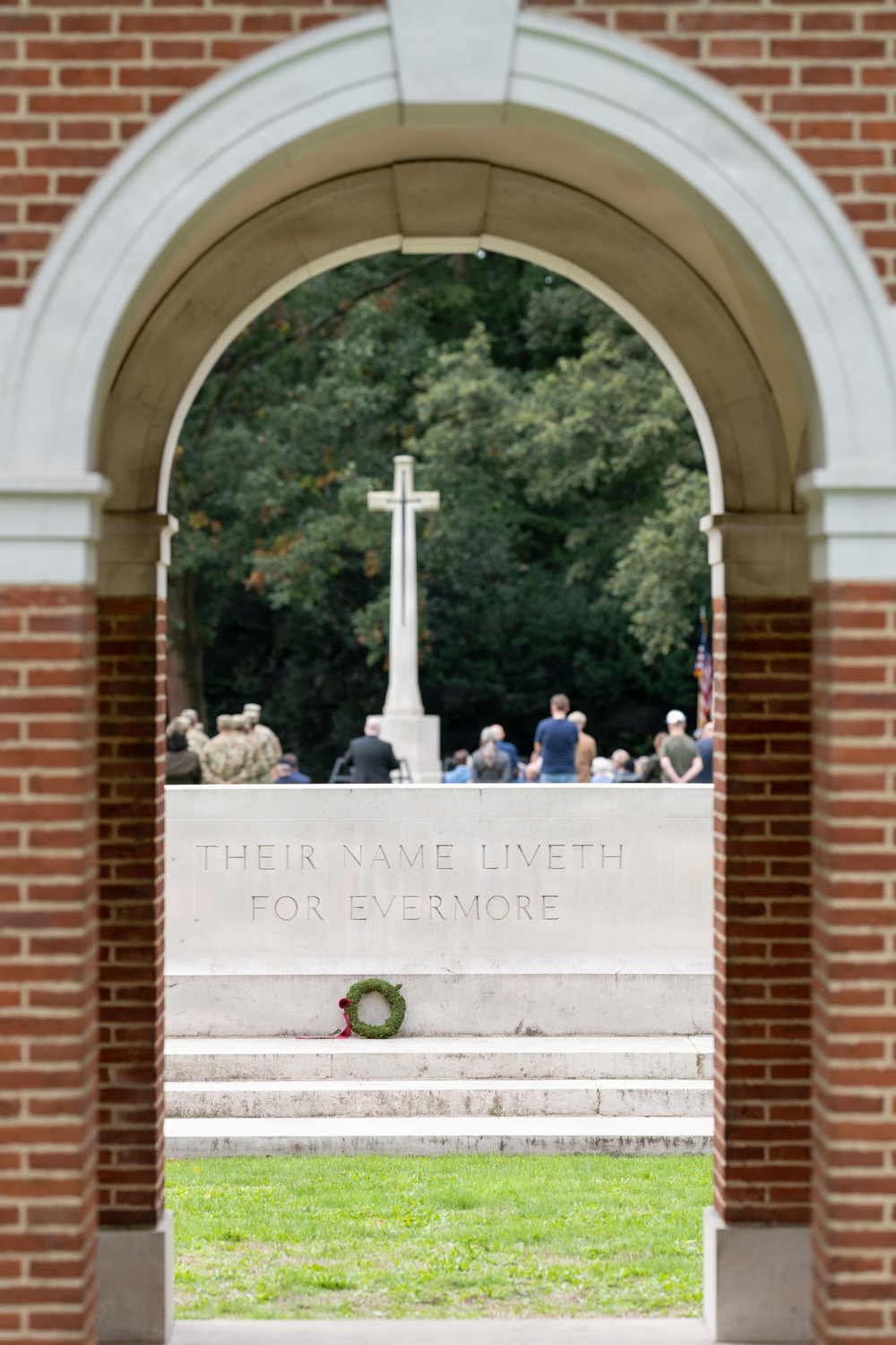 The image shows a memorial ceremony viewed through a brick archway at what appears to be a Commonwealth War Graves Commission cemetery. A stone monument bears the inscription 'THEIR NAME LIVETH FOR EVERMORE,' a phrase from the Book of Sirach selected by Rudyard Kipling for CWGC memorials. A small green wreath with a red ribbon has been placed at the base of the monument, and a group of attendees faces a large Cross of Sacrifice in the background.