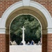 The image shows a memorial ceremony viewed through a brick archway at what appears to be a Commonwealth War Graves Commission cemetery. A stone monument bears the inscription 'THEIR NAME LIVETH FOR EVERMORE,' a phrase from the Book of Sirach selected by Rudyard Kipling for CWGC memorials. A small green wreath with a red ribbon has been placed at the base of the monument, and a group of attendees faces a large Cross of Sacrifice in the background.