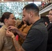 A military promotion or graduation ceremony is taking place indoors, likely at a naval facility. A man with visible tattoos is pinning an insignia or device onto the collar of a female sailor in a tan/khaki uniform. Family members or guests are observing the pinning ceremony in the background.