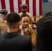 A military promotion ceremony is taking place, likely in the United States Navy, where a female sailor in khaki uniform is having insignia or rank devices pinned onto her collar by two civilians, presumably family members. Two senior enlisted Navy chiefs in khaki uniforms and combination covers stand in the background before a large American flag. The scene reflects the traditional Navy pinning ceremony marking a promotion in rank.