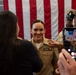A U.S. Navy service member in dress khaki uniform is being congratulated or having insignia pinned during what appears to be a promotion or advancement ceremony. An American flag is displayed prominently in the background. Attendees are capturing the moment on smartphones.