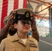A U.S. Navy chief petty officer promotion ceremony is taking place, with a service member having the chief's combination cover (hat) placed on their head by another individual. The subject is wearing the tan Service Khaki uniform with gold insignia, ribbons, and a warfare device. A large American flag is displayed prominently in the background of what appears to be a hangar or large indoor military facility.