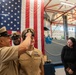 A U.S. Navy promotion or advancement ceremony is taking place inside what appears to be a military facility or gymnasium. A senior enlisted sailor is placing a chief petty officer cover (hat) on a female sailor's head, indicating a pinning or advancement ceremony. Two civilian women observe and smile in the background, alongside another uniformed sailor.