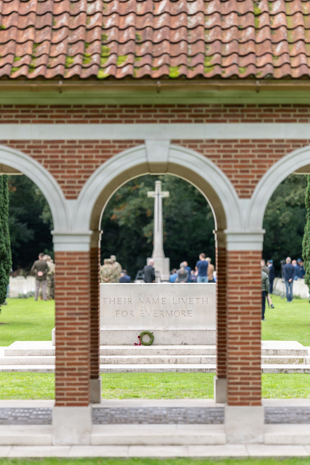 A Commonwealth War Graves Commission cemetery is shown through a brick archway, featuring the Stone of Remembrance inscribed with 'THEIR NAME LIVETH FOR EVERMORE.' A commemorative ceremony is taking place in the background, with a group of civilians and military personnel gathered around the Cross of Sacrifice. A small wreath with red poppies has been placed on the steps of the Stone of Remembrance.
