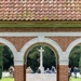A Commonwealth War Graves Commission cemetery is shown through a brick archway, featuring the Stone of Remembrance inscribed with 'THEIR NAME LIVETH FOR EVERMORE.' A commemorative ceremony is taking place in the background, with a group of civilians and military personnel gathered around the Cross of Sacrifice. A small wreath with red poppies has been placed on the steps of the Stone of Remembrance.