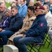A memorial ceremony is taking place at what appears to be a military cemetery, with rows of white grave markers visible in the foreground. Attendees are seated on folding chairs, including a man wearing an 82nd Airborne Division jacket and maroon beret prominently in the foreground. An active-duty soldier in Army Combat Uniform with Ranger and 7th Infantry Division patches is visible on the left edge.