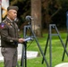 A U.S. Army officer in dress uniform is delivering a speech at a podium with a microphone, appearing to be at a military memorial ceremony. The setting appears to be a military cemetery or memorial ground, with grave markers visible in the foreground and background. Several easels are arranged in the background, likely holding commemorative photographs or plaques.