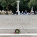 A Commonwealth War Graves Commission memorial stone bears the inscription 'THEIR NAME LIVETH FOR EVERMORE', a phrase selected by Rudyard Kipling. A small moss wreath with red poppies rests on the stone's base steps. In the background, a group of military personnel and civilians are gathered near a large stone cross for what appears to be a remembrance ceremony.