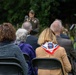 An outdoor military ceremony or memorial event with civilians and military personnel seated in folding chairs facing a uniformed soldier at a podium. A woman in the foreground wears a red, white, and blue scarf bearing the 82nd Airborne Division patch ('AA' insignia). The setting appears to be a wooded or garden area, suggesting a commemorative or remembrance event.