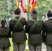 A U.S. Army color guard of four soldiers in olive dress uniforms is participating in a formal military ceremony, viewed from behind. Two center soldiers carry flags, including a visible U.S. Army flag and what appears to be an American flag, while the two outer soldiers carry rifles. A crowd of attendees and a monument or memorial structure are visible in the background.
