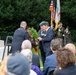 A formal wreath-laying ceremony is taking place at what appears to be a war memorial or monument. Two men in formal attire, one wearing military medals and a blue beret, are presenting a floral wreath to an official in a decorated uniform. A color guard stands in the background holding an American flag and what appears to be a unit or organizational flag, while seated attendees observe the ceremony.
