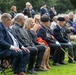 A formal outdoor memorial or commemorative ceremony attended by a large crowd seated on chairs on a grass lawn. Several individuals wearing military uniforms with medals and berets are prominently visible in the foreground, suggesting this is a veterans' remembrance event. White grave markers or headstones are faintly visible in the background, indicating the setting is likely a military cemetery.