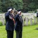 Two elderly veterans in dark blazers and black berets are saluting during a memorial ceremony at a military cemetery with rows of white Commonwealth-style headstones. Both men wear service medals and white gloves, with one displaying a red sash and a visible poppy. Several other attendees, including a person in military uniform and a civilian photographing the event, are visible in the background.