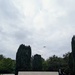 A Commonwealth War Graves Commission memorial stone inscribed with 'THEIR NAME LIVETH FOR EVERMORE' is prominently displayed in the foreground of a military cemetery. A wreath has been laid at the base of the stone, and a small group of formally dressed attendees can be seen in the background near a white cross. Two aircraft are visible flying in formation overhead, suggesting an active commemorative ceremony.