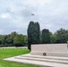 A Commonwealth War Graves Commission cemetery is shown, featuring the iconic Stone of Remembrance inscribed with 'THEIR NAME LIVETH FOR EVERMORE' — a phrase chosen by Rudyard Kipling. A wreath has been laid at the base of the stone, and a military aircraft is visible flying overhead, suggesting a commemorative ceremony is taking place. Rows of uniform white headstones are visible in the background among well-maintained green lawns.