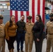 A group of six people pose for a formal photograph in front of a large American flag in what appears to be a military facility or hangar. Three individuals are wearing U.S. Navy uniforms, including one in a dress uniform with ribbons and a cover (cap), while the other three are in civilian attire. The setting suggests a military ceremony or promotion event.