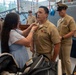 A military promotion or pinning ceremony is taking place inside a facility associated with the USS Whitehat. A woman in a light blue dress is pinning insignia onto a U.S. Navy Chief Petty Officer's uniform collar, a traditional part of Navy chief pinning ceremonies. Several other uniformed Navy personnel are present in the background observing the event.