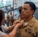 A military promotion or pinning ceremony is taking place, likely in a U.S. Navy context. A woman is pinning a new insignia or rank device onto the collar of a male sailor in khaki service uniform. The setting appears to be inside a naval museum or ceremonial hall, with other uniformed personnel visible in the background.