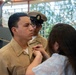 A military promotion or pinning ceremony is taking place, likely in the U.S. Navy. A woman in civilian clothing is pinning a new insignia or rank device onto the collar/chest of a sailor in khaki service uniform. A second sailor wearing a chief petty officer's combination cover observes in the background.