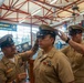 A U.S. Navy promotion or advancement ceremony is taking place inside a military facility. Two senior Navy personnel are placing a chief petty officer cover (hat) on a newly advanced sailor, a traditional part of the CPO pinning/advancement ceremony. The setting features Navy banners and signage, including 'America's Navy: A Global Force for Good.'