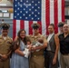A group of seven people pose for a formal photograph in front of a large American flag in what appears to be a naval facility or ship interior. Three individuals are wearing U.S. Navy uniforms, including khaki service uniforms with combination covers, while the others are in civilian attire. A newborn baby dressed in a miniature sailor outfit is being held by the central uniformed sailor, suggesting a celebratory family occasion.