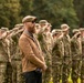 A civilian man with a red beard, wearing a tan jacket, flat cap, and sunglasses, stands in front of a large formation of military personnel in camouflage uniforms who are saluting. The setting appears to be an outdoor military ceremony on a grassy field with trees in the background. A partially visible woman with blonde hair is seen on the right edge of the frame.