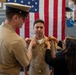 A U.S. Navy promotion ceremony is taking place in what appears to be a hangar bay or indoor military facility. Two individuals — a senior uniformed officer and a civilian woman — are simultaneously pinning new rank insignia onto the collar of a sailor being promoted. An American flag is prominently displayed in the background.