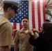 A military promotion ceremony is taking place in front of a large American flag. A woman is pinning new insignia onto the collar of a U.S. Navy sailor in tan/khaki service uniform, while a senior officer in a cover (hat) observes. The setting appears to be an indoor military facility or hangar bay.
