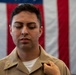 A young male U.S. Navy sailor in a tan/khaki uniform stands before an American flag with eyes closed while another person's hand pins a gold insignia onto his collar. The scene appears to depict a military promotion or pinning ceremony. Two gold chief petty officer anchors are visible on the collar points of his uniform.