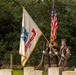 A U.S. Army color guard unit is presenting colors at what appears to be a military cemetery ceremony. Four soldiers in Army Service Uniforms are carrying the United States Army flag (established 1775) and the American flag, while two others hold rifles at attention. The setting among grave markers and lush green trees suggests a formal memorial or commemorative event.