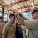 A military promotion or pinning ceremony is taking place inside what appears to be a naval facility or hangar. A senior naval officer with visible tattoos on his forearm is placing or adjusting a cover (hat) or insignia on a junior sailor's head, while other uniformed personnel and a civilian woman observe. The setting features a distinctive red steel truss ceiling structure.