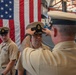 A U.S. Navy promotion or advancement ceremony is taking place inside what appears to be a naval hangar or facility. A senior enlisted sailor is placing a chief petty officer combination cover (hat) on a newly advanced sailor, while another chief petty officer stands at attention in the background. A large American flag serves as a ceremonial backdrop.
