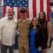 A U.S. Navy service member in dress khaki uniform poses with three civilians in front of a large American flag, likely at a military ceremony or promotion event. The setting appears to be inside a naval facility or museum, with what appears to be a ship or vessel visible in the background. The group is smiling, suggesting a celebratory occasion.