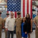 A group of seven people pose for a formal photograph in front of a large American flag displayed in what appears to be a naval facility or museum. Four individuals are wearing U.S. Navy uniforms with chief petty officer covers, while three civilians stand among them, suggesting a military ceremony or promotion event. The setting includes naval equipment and decorative signal flags in the background.