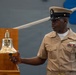 A U.S. Navy enlisted sailor in khaki service uniform is ringing or holding a polished brass ship's bell mounted on a wooden stand. The bell is engraved with 'US' and appears to be part of a formal naval ceremony. The setting appears to be aboard a naval vessel or in a naval facility.