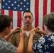 A U.S. Navy promotion ceremony is taking place in what appears to be a ship's hangar bay or military facility. Two women are pinning new rank insignia onto the collar of a male sailor in a tan/khaki dress uniform, a traditional part of Navy promotion ceremonies. An American flag serves as the backdrop, and another uniformed sailor is visible in the background.
