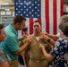 A U.S. Navy promotion ceremony is taking place in what appears to be a naval facility or museum, with an American flag as a backdrop. Family members and colleagues are pinning new rank insignia onto a sailor's uniform collar, a traditional part of military promotion ceremonies. A second uniformed Navy sailor wearing a combination cover (hat) is visible in the background.