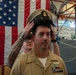A U.S. Navy Chief Petty Officer promotion ceremony is taking place in what appears to be a ship's hangar bay or naval facility. A person behind the newly promoted sailor is placing the distinctive khaki combination cover (cap) with a Chief Petty Officer device onto his head, a traditional part of the CPO pinning/frocking ceremony. A large American flag serves as the backdrop for the formal military occasion.