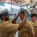 A U.S. Navy promotion ceremony is taking place inside what appears to be a naval facility or recruiting station. A senior enlisted sailor is placing a chief petty officer cover (hat) on a newly promoted sailor, a traditional part of the CPO pinning/frocking ceremony. Several other sailors and civilians are present as witnesses to the event.