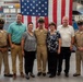 A group of seven people pose for a formal photograph in front of a large American flag in what appears to be a military facility or naval training center. Three individuals are wearing U.S. Navy uniforms, including khaki service uniforms and a dress uniform with a combination cover, suggesting this is a military ceremony or promotion event. The remaining four individuals appear to be civilian family members or guests.
