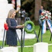 A woman in a navy blue polka dot dress is delivering a speech or reading at what appears to be a military memorial ceremony. She stands at a microphone podium surrounded by commemorative wreaths on stands, with grave markers visible in the foreground. The setting is an outdoor military cemetery or memorial garden.