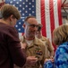 A U.S. Navy officer in khaki service uniform is having insignia or medals pinned onto his uniform by a woman, likely a family member, during what appears to be a military promotion or retirement ceremony. A large American flag is displayed prominently in the background, and a child in a blue jacket is also participating in the pinning. A second uniformed service member is visible in the background.